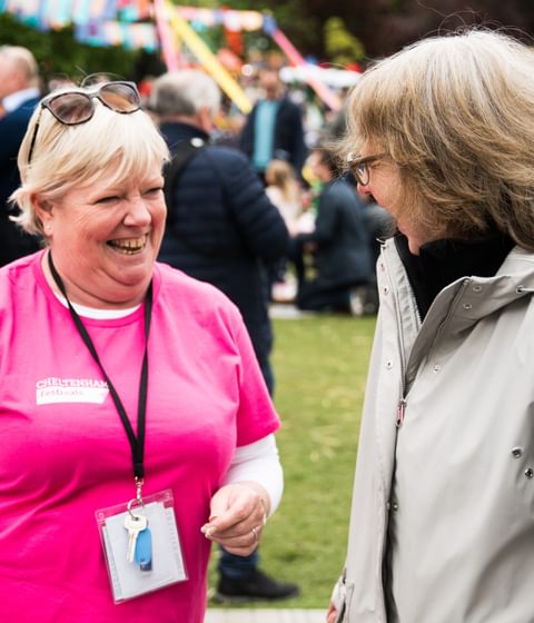 A woman wearing a pink t-shirt talking to a Festival-goer. She is smiling.