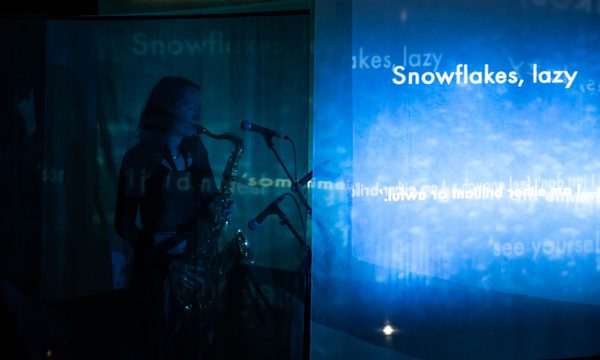 A woman plays the saxophone in the dark. Projected on a screen next to her are the words 'snowflakes, lazy'.