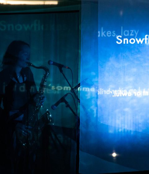 A woman plays the saxophone in the dark. Projected on a screen next to her are the words 'snowflakes, lazy'.