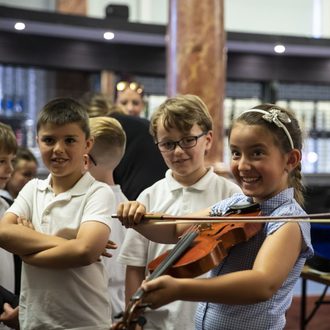 Primary school aged girl playing the violin and smiling with two primary school aged boys in the background listening