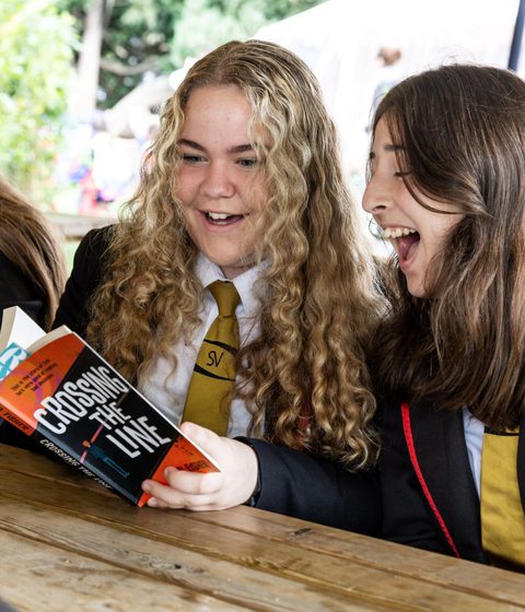 Two school children laughing while reading Crossing the Line in the Picnic Area at Cheltenham Literature Festival.