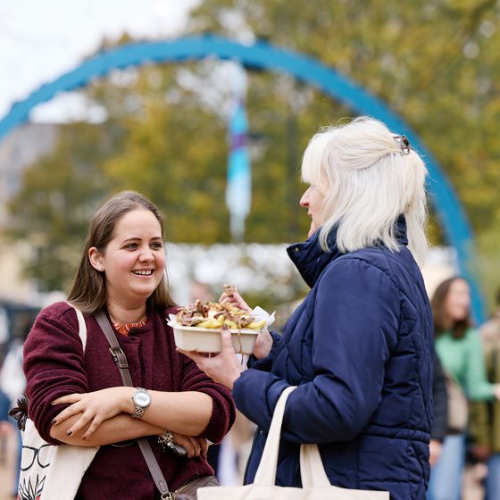 Two women stood under the Cheltenham Literature Festival arch eating food