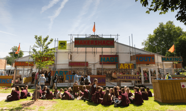 A photo of the Makershack, a tent with rustic looking signage, with a crowd of people sat in front of it in the sun.